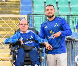 Two men in blue sports jerseys stand by a fence at a stadium, one in a wheelchair smiling with a teammate beside him.