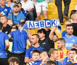 Crowd of soccer fans in blue jerseys cheering in stadium stands, with a blue banner held up by several supporters in the center-left.
