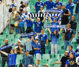 Crowd of blue-clad soccer fans in stadium stands waving striped scarves and cheering sunlit green seats behind them