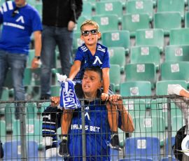 Young boy in a blue jersey and sunglasses sits on an adult’s shoulders, cheering at a stadium behind a safety barrier.