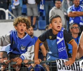 Two young boys lean over a stadium railing, cheering with mouths open in blue team jerseys as a crowd watches from the stands behind them.