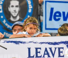 Young girl with a purple headband leaning over a railing at a crowded event, a blue banner behind her.