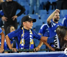 Group of young fans in blue jerseys leaning on a railing at a stadium, cheering during a game.